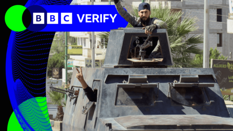 A fighter leans out of a car in Syria. He is holding his arm aloft and leaning on a machine gun.