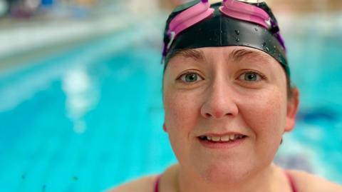 Gemma Stevenson is looking directly at the camera and smiling slightly while wearing a black swimming cap and pink goggles, which are off her eyes and resting on her head. She is holding onto the side of the pool.  Behind her the water is a bright blue and there are no other people in the swimming pool. There are droplets of water on her nose and the edges of her swimming cap. The straps of her swimming costume are pink and can be seen on shoulders, just in frame of the camera. 