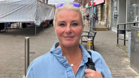 Julie Harriman, who has blonde hair which has been tied up and brown eyes. She is wearing a denim shirt and smiling in the photo. Behind her an empty shop unit can be seen.
