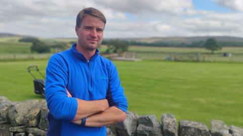 Farmer James Jackson is pictured with arms folded in a blue fleece top, with a field and dry-stone wall behind him