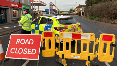 A road closure sign, police car and a police officer standing near some police tape at the foreground of the image. In the background, the A38 stretches out, with an ESSO garage on the left and some more police vehicles in the background. 