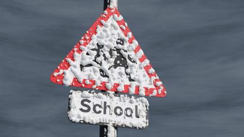 A school road sign on a pole covered in snow with a dark grey cloudy sky behind