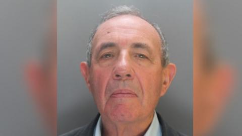A mugshot of John Allen. He wearing dark jacket and shirt and standing against a grey background. 