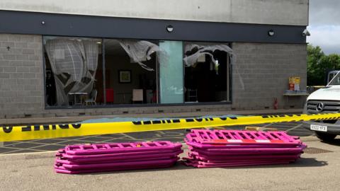 The front of the building suffered smashed windows and ripped curtains. Some furniture is partially visible inside the hotel. Pink barriers are stacked in front of disabled parking bays and a white van can be seen parked on the right. A yellow police cordon stretches from one end to the other.