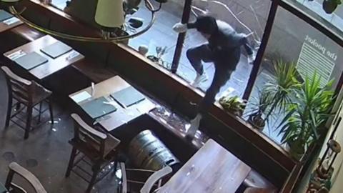 CCTV footage shows a recent break-in at The Pocket coffee shop in Belfast city centre. A male in a tracksuit has just smashed the glass windows with a keg, pictured on the floor. He is climbing into the building, there are numerous tables chairs and plants visible in the restaurant.