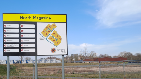 A sign denotes the firms on the enterprise zone. It stands in front of wasteland ready for development. The words North Magazine are in black font on a yellow background at the top of the sign.