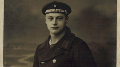 A black and white portrait of a young man in a naval uniform. He has a heavy coat on with brass buttons, and a navy smock underneath, as well as a cap.