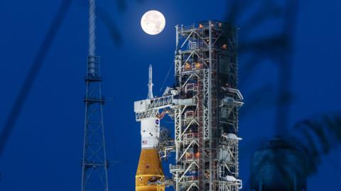 A large and very bright full Moon appears directly on the top of Nasa's Space Launch System rocket at night. The orange and white rocket is sitting next to a steel support structure called a mobile launcher.
