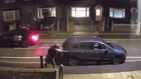 A man in a grey looking coat with the hood raised, carrying a black backpack, stands on a residential street holding his right arm straight forward. In the background is a dark Mercedes car with its brake light illuminated red and the rear passenger side door open. 