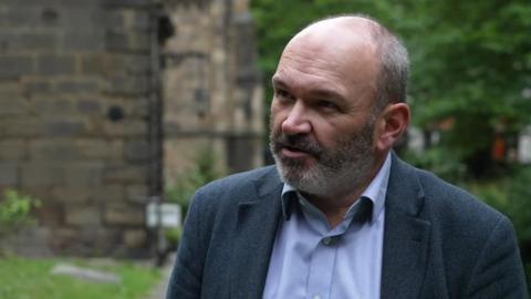 Head and shoulder shot of a man with thinning, cropped grey hair and a closely cropped grey beard. He is wearing a dark blue jacket and light blue shirt, open at the collar.