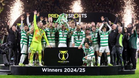 Celtic wear green and white striped tops. They are cheering as pyrotechnics go off behind them. They're lifting the trophy standing on a platform which says: "Winners 2024/2025"