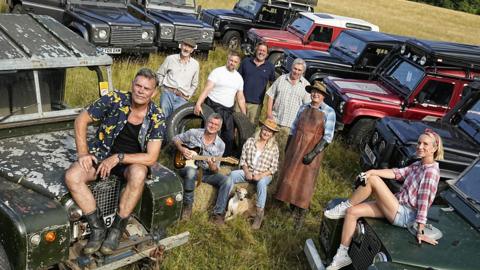 A group of people are posing for a photo with a gathering of parked land rovers in a field. A man and women at the front of the image are sat on the bonets of two of the land rovers. The rest are sat and stood around a pile a hay bales.