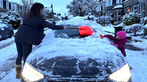 A mother and daughter clear the snow-laden windscreen of a car in the early hours. The car is on an upmarket residential street with sandstone properties, trees and bushes.