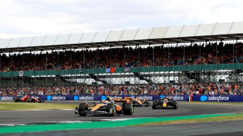 Formula 1 cars racing during the British Grand Prix at Silverstone. Cars from McLaren, Red Bull and Ferrari are all shown against the backdrop of a packed grandstand.
