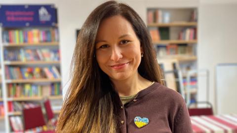 Iryna has long brunette hair. She is smiling and has a light brown jumper on and is wearing a heart-shaped badge with the yellow and blue of the Ukrainian flag.