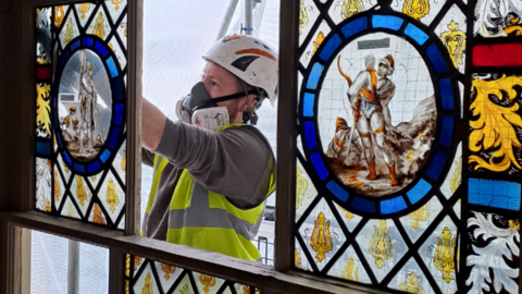 A man wearing a fluorescent hi-vis jacket and helmet is working on a stained glass window. The window is at the top of a staircase and the man is outside the building on scaffolding.