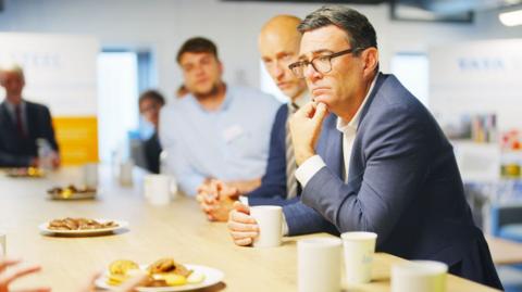 Andy Burnham with Stephen Kinnock sat at a table with bicsuits and mugs. They both have blue suits on. They are in focus, while other people are also sat there who are blurred.