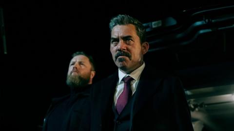 Two men are looking forward. It's dark.
The man in the foreground has greying short hair and a moustache.
He is wearing a dark coat, over a dark suit. His shirt is white and his tie purple.
The man behind is dressed fully in black. He has lighter hair and a full beard