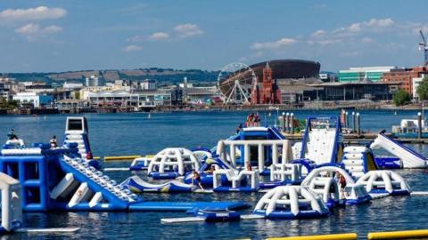 A blue and white inflatable assault course on water with the iconic Cardiff Bay skyline, including a big wheel, the Wales Millennium Centre  and the Pierhead building, in the background.