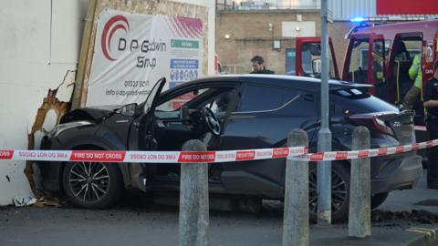 A black suv crossover has hit hoardings on the side of a white building. There is 'Fire do not cross' tape wrapped round the scene. 