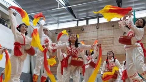 A collection of dancers wearing white and red clothes holding red and yellow cloths 