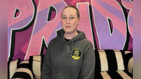 CEO of Pie Factory Music, Zoë Carassik, wearing a grey hoodie and stood against the backdrop of the Pie Music Factory charity headquarters in Ramsgate. 'Pride' is painted in pink and purple on the wall and Zoe is stood in front of a black and white striped sofa.