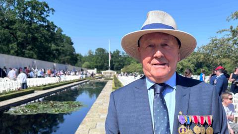 A man looking at the camera, wearing a straw hat and smart navy suit and tie. He has five medals pinned to the suit. Behind him is a pool of water, which is part of the cemetery's memorial garden, and several rows of white plastic chairs with people sitting in some of them. 