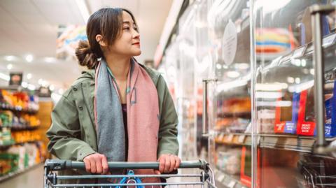 A woman holding a trolley in a supermarket looking to refrigerated shelves.