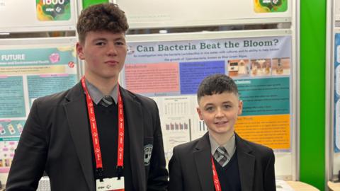 Two schoolboys wearing school uniforms pose for a picture in front of a display. Both have brown hair and are wearing a black blazer and a grey shirt with a black, and blue stripped tie. Posters about blooms of bacteria are behind them. They're wearing red lanyards. 