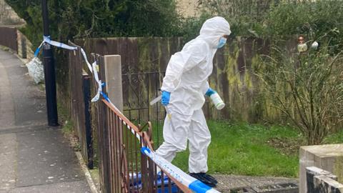 A police forensics officer wearing full coveralls and a mask walks away from a metal fence covered in police tape along a path with a shrub to the right.