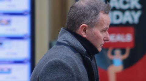 Gary Marsden walking down the street. He looks straight ahead of him, has a neutral expression on his face, and wears a blue/grey woollen coat with the collar raised.