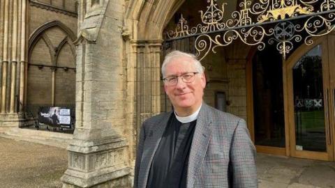 The Reverend Tim Alban Jones wearing a checked jacket and dog collar, smiling for the camera outside the cathedral