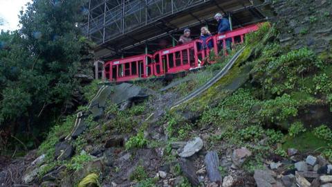A landslip on a road in Dartmouth. There are red plastic fence barriers at the top of the landslip, where there are three people stood talking.