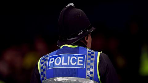 A police officer, wearing a traditional hat and a hi-vis vest, stands with his back to the camera against a blurred black background