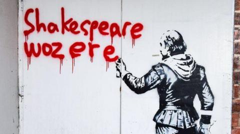 Art painted on white double doors on a brick building. It is an image in black of a character that looks like William Shakespeare from behind. He is holding a spray can, and the words "Shakespeare woz ere" have been written in red ink.