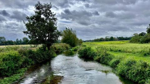 A body of water sits between two green fields under dark grey and black clouds