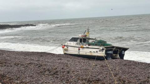 A small white fishing boat with orange buoy attached to it, washed up on a pebbly beach with the waves crashing on to it.