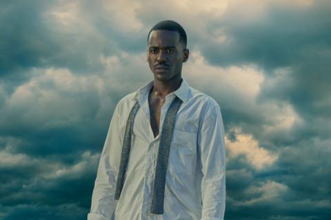 Colour image of Ncuti Gatwa wearing white shirt against backdrop of storm clouds
