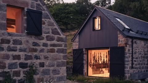 Two stone buildings face each other's gable ends as it gets dark. Warm light floods out of the studio's large window