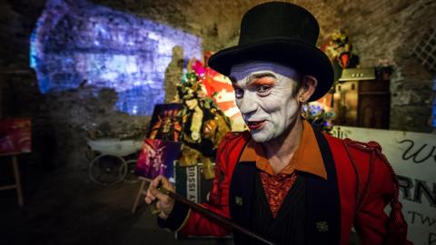 Doug wearing white face make up and costume and a hat. Doug is holding a cane and looks like he is in mid-conversation with the photographer and performing while smiling. It looks like he is standing in a cave. There are circus props behind him.