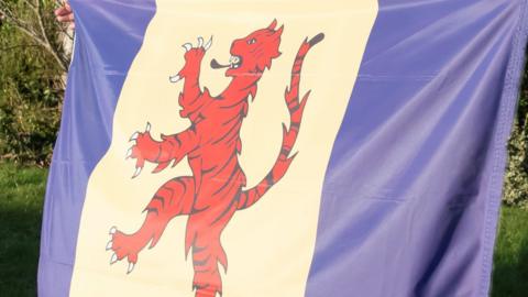 Close-up view of the Fenland flag. The flag has two vertical strips of blue at each end of the flag, separated by a strip of white in the centre. On the white strip is an image of a tiger.