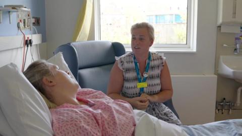 A woman in a hospital bed, with a nurse sat in the chair by her bedside talking to her.