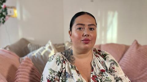 Suzannah Mahe is sat on a pink sofa at home wearing a floral dress and looking into the camera.