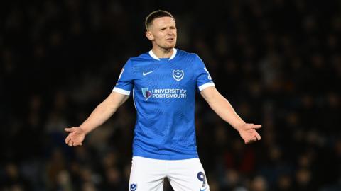 Colby Bishop holding out his arms in frustration while wearing a blue Portsmouth home kit and white shorts with the number 9 and the club badge printed on