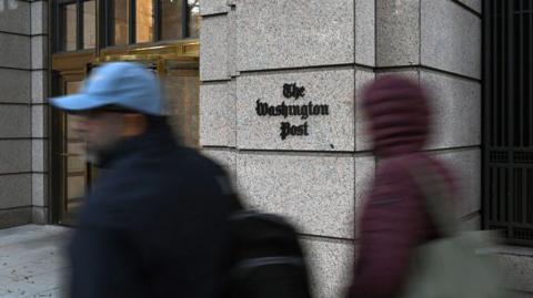 People walk by the outside of a building that has a sign that reads "The Washington Post"