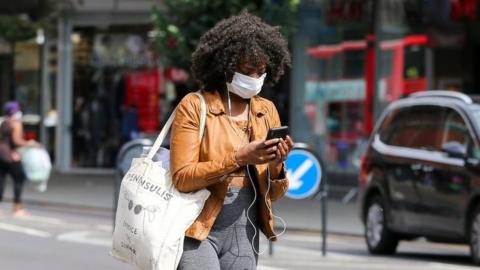 A woman wearing a face mask is seen using a mobile phone while walking on the street.