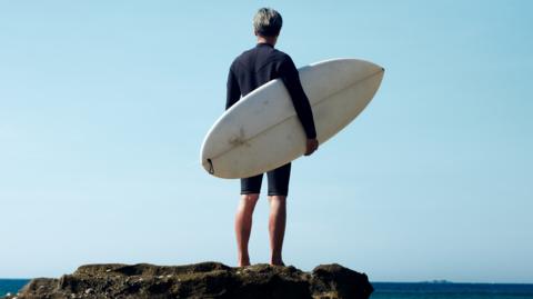 A man wearing a wetsuit and carrying a surfboard stands on a rock near the ocean