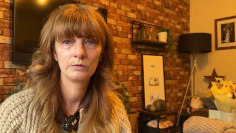 A woman with ginger hair and a fringe, she has dark blue eyes and glasses on her head. She looks upset and is staring into the camera. She is sitting in a living room, with a TV on the wall behind her and furniture to the right.