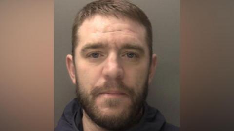 Billy Ormiston - a custody image of a man with short brown hair, a moustache and short beard. He is wearing a dark blue hoody and stood against a grey background