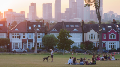 With the high-rises of Nine Elms in the distance, people sit in together in Ruskin Park while a solstice sun sets over period homes behind them 